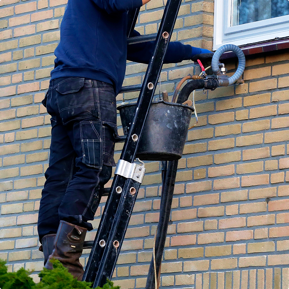 Installer on a ladder removing cavity wall insulation from a brick house with a hose